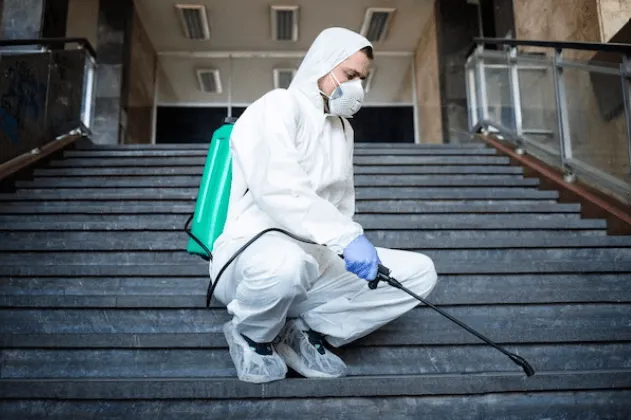 Un disinfestatore, accuratamente coperto, mentre usa il nebulizzatore sulle scale di entrata di un condominio